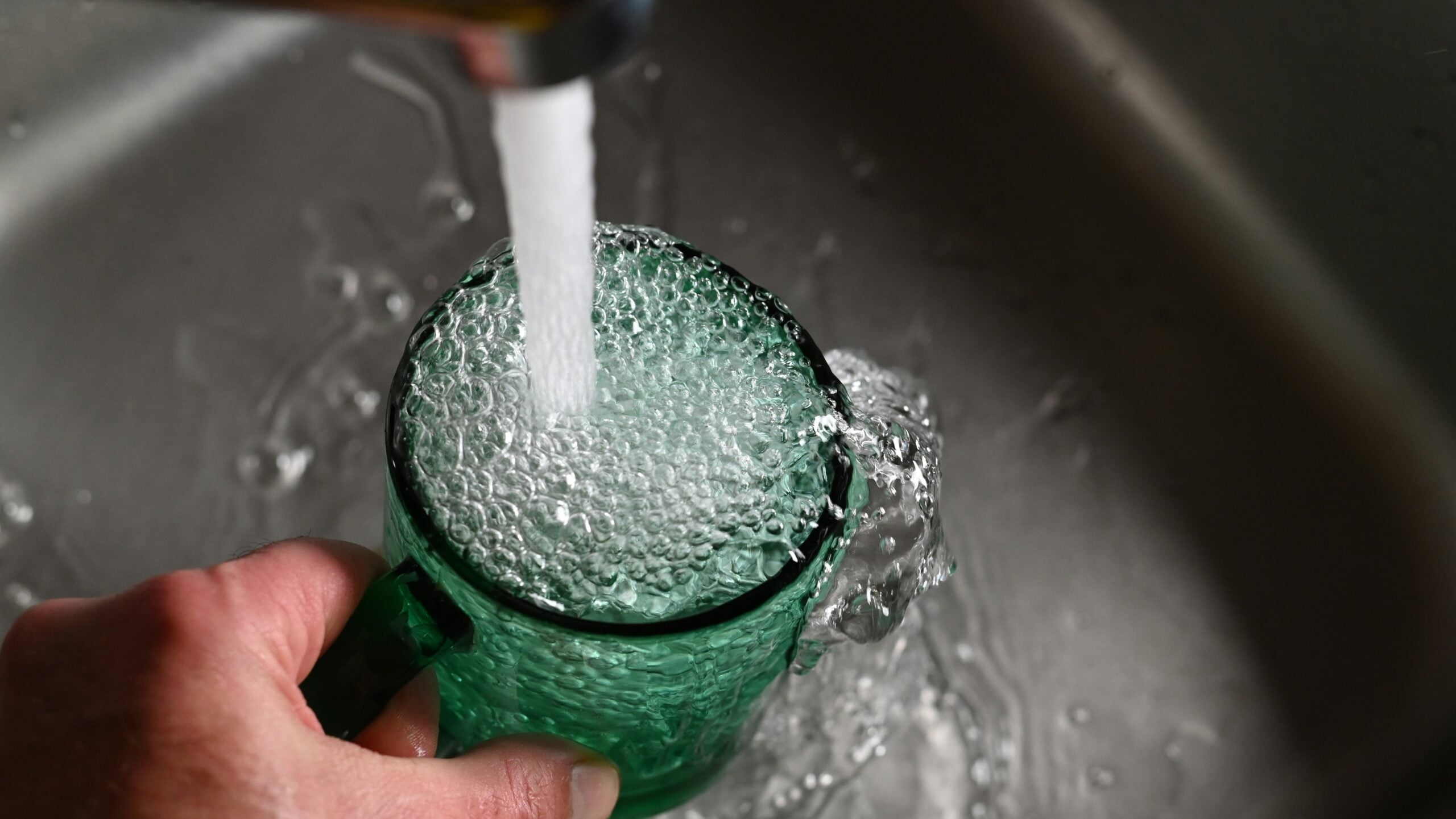 Water overflowing a green glass mug held under a faucet in a steel sink.